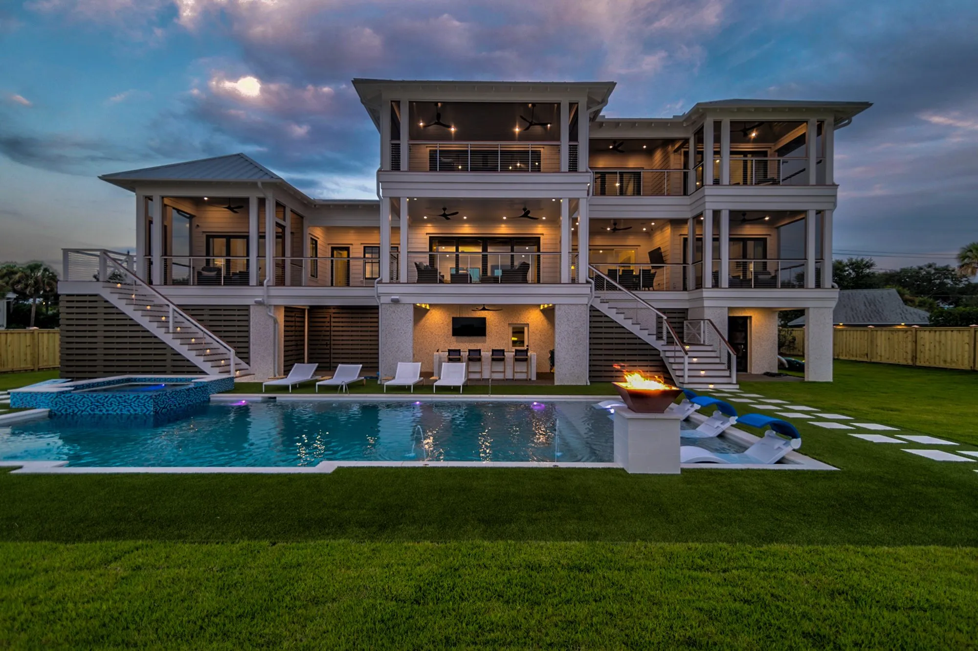 Three-story waterfront home with a lit pool and fire bowls at twilight